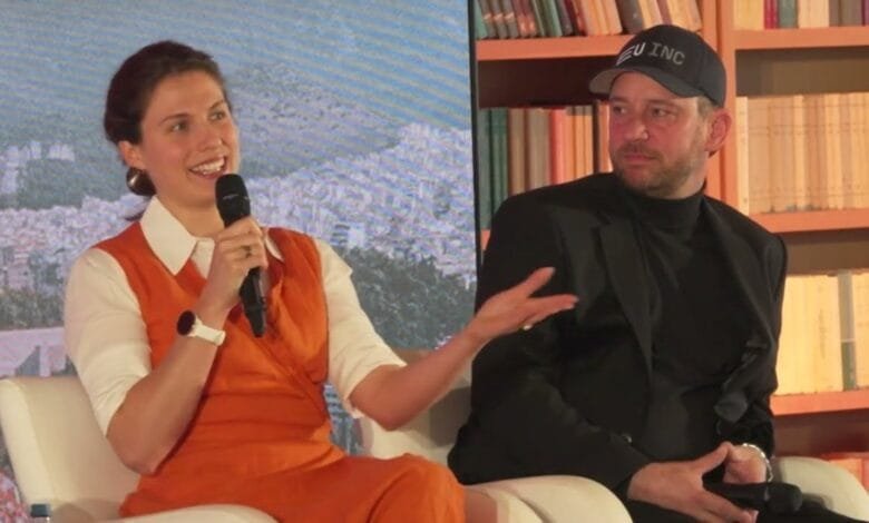 A woman in an orange dress and a man in a black jacket participate in a panel discussion. A scenic backdrop is visible.