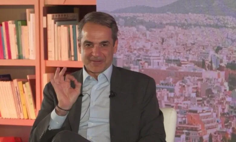 Kyriakos Mitsotakis, Prime Minister of Greece, sits for an interview, giving an 'OK' hand gesture. A cityscape backdrop is visible.