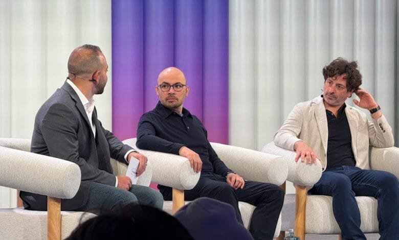 Three men sit in light gray armchairs on a stage, engaged in conversation. A purple backdrop is visible.