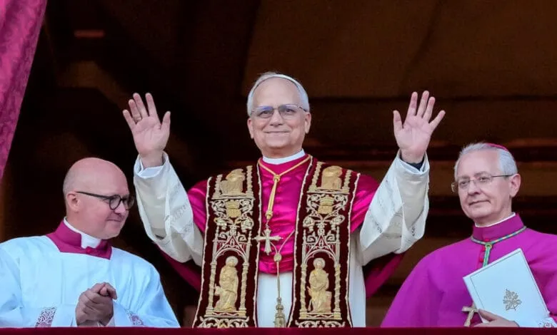 Pope Francis waves to the crowd from a balcony, flanked by two other clergy members in ceremonial robes.