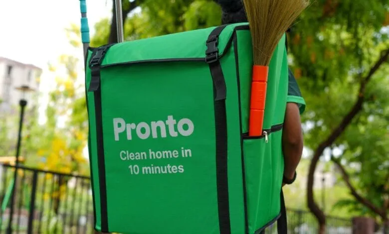 A green Pronto cleaning service backpack with a broom attached, worn by a person outdoors.
