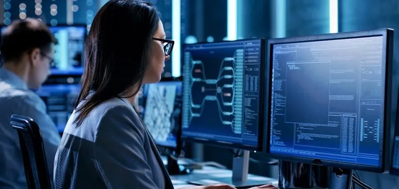 Woman in glasses intently works at multiple computer monitors displaying complex data and code in a dimly lit tech environment.