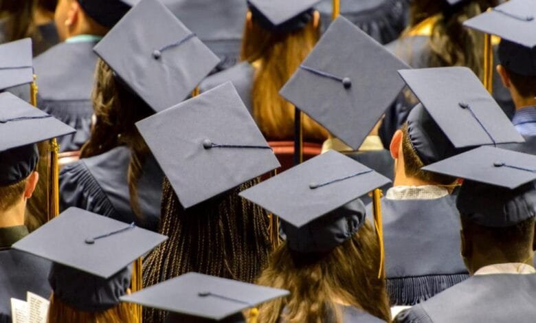 Many graduates in navy blue caps and gowns sit together, viewed from behind.