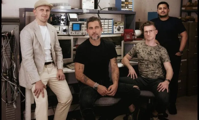 Four men pose in a vintage electronics workshop. They're seated and standing amidst old computers and equipment.