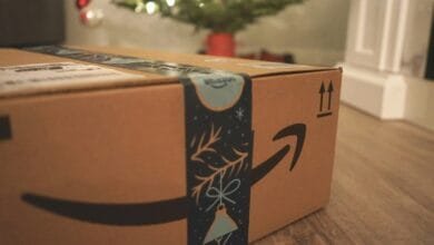 Close-up of an Amazon package with a festive holiday design on the tape, sitting on a floor near a blurred Christmas tree.