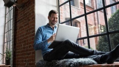 Man sits on window seat, legs outstretched, using a laptop. He smiles, looking at the screen in a relaxed setting.