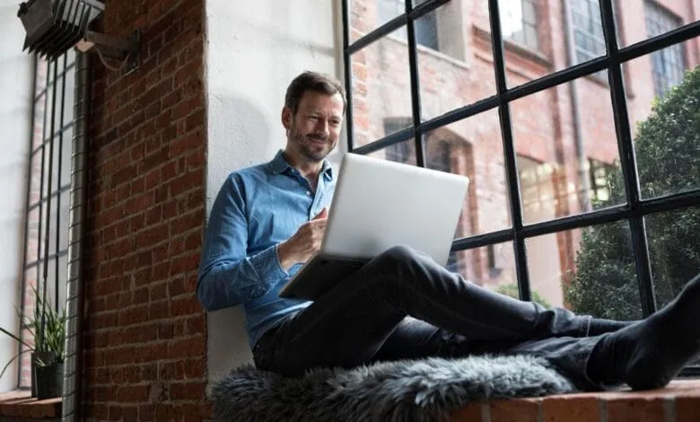 Man sits on window seat, legs outstretched, using a laptop. He smiles, looking at the screen in a relaxed setting.