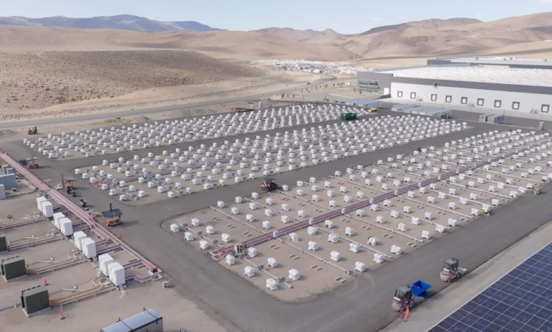 Aerial view of a large-scale battery energy storage system with numerous white battery packs arranged in rows, next to a solar farm and warehouse.