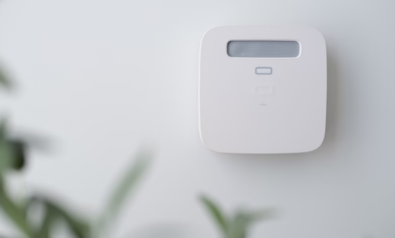 A small, square, white smart home device is mounted on a white wall. Green plant leaves are blurred in the foreground.