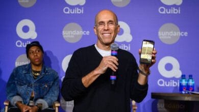 Jeffrey Katzenberg, Quibi founder, holds a phone showcasing the app's interface at Sundance 2020. Two other individuals sit beside him.