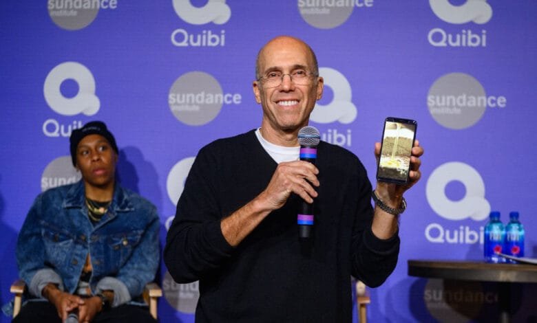 Jeffrey Katzenberg, Quibi founder, holds a phone showcasing the app's interface at Sundance 2020. Two other individuals sit beside him.