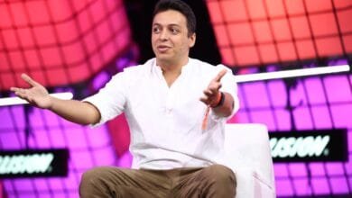 A man in a white shirt and brown pants sits on a white chair, gesturing with open hands. He's speaking at a conference with a purple and red background.