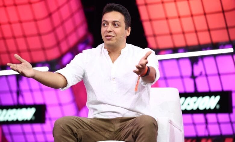 A man in a white shirt and brown pants sits on a white chair, gesturing with open hands. He's speaking at a conference with a purple and red background.