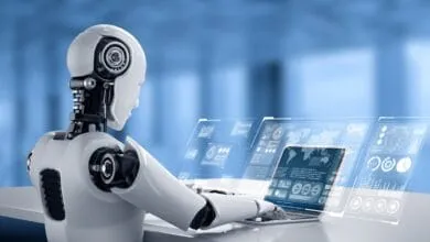 A white robot sits at a desk using a laptop with holographic displays showing data charts and graphs.