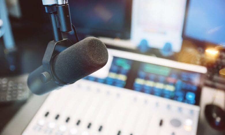Close-up of a microphone in a recording studio, with a soundboard and computer monitors in the background.