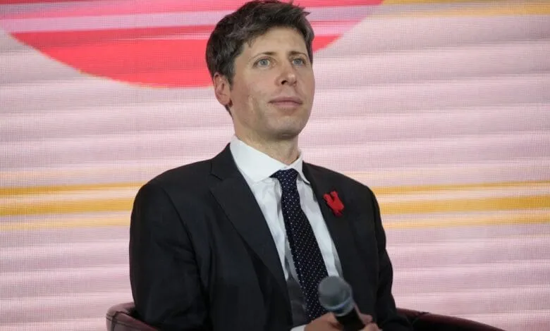 Sam Altman, CEO of OpenAI, sits on stage holding a microphone, looking thoughtfully upward. He's wearing a dark suit and patterned tie.
