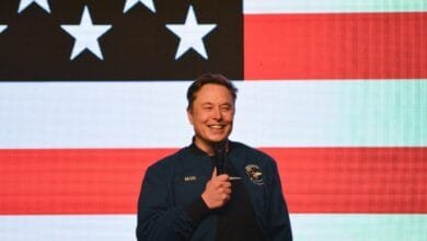 Elon Musk, CEO of SpaceX and Tesla, smiles while speaking at an event with an American flag backdrop.