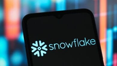 Close-up of a smartphone displaying the Snowflake logo and company name against a blurred background of stock market charts.