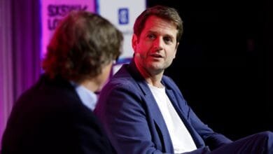 A man in a blue blazer speaks at a conference, another person is partially visible to the left.