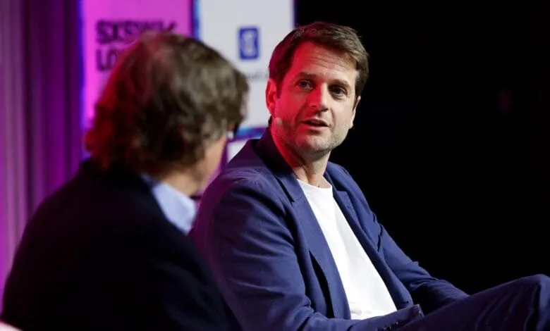 A man in a blue blazer speaks at a conference, another person is partially visible to the left.