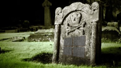 An old, weathered gravestone in a graveyard at night features a skull and crossbones carving and a four-paneled design resembling a window.
