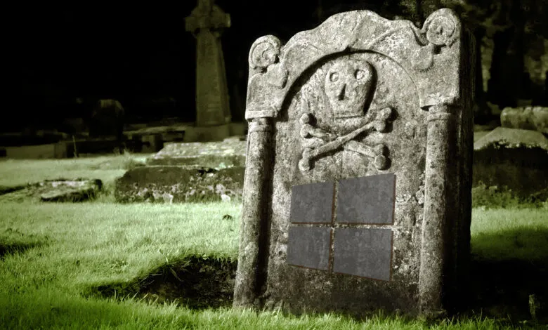 An old, weathered gravestone in a graveyard at night features a skull and crossbones carving and a four-paneled design resembling a window.