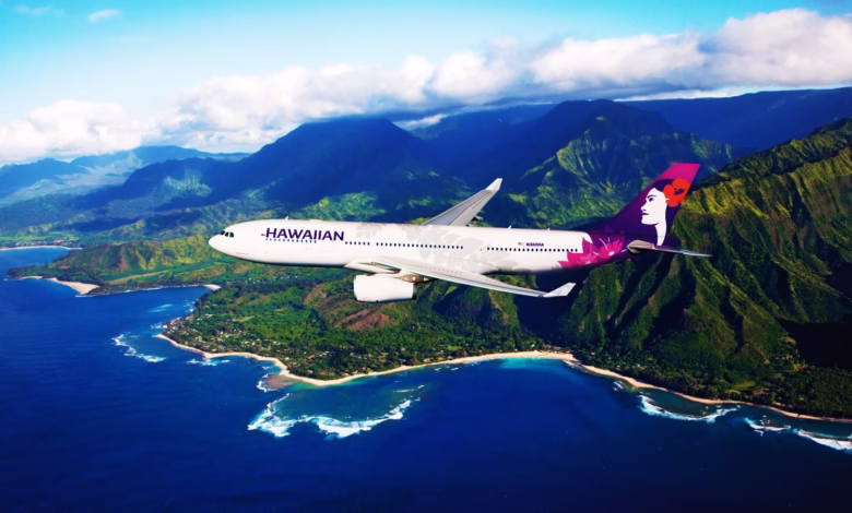 Hawaiian Airlines plane flies over lush green mountains and turquoise ocean in Hawaii.