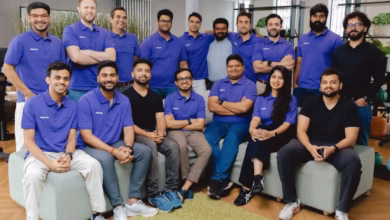 A diverse team of 16 people, mostly men, smiles for a group photo. They wear purple polo shirts and sit or stand on a light green couch and floor.