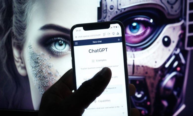 Close-up of a hand holding a smartphone displaying the ChatGPT interface. A blurry background shows a stylized female face and a robotic figure.