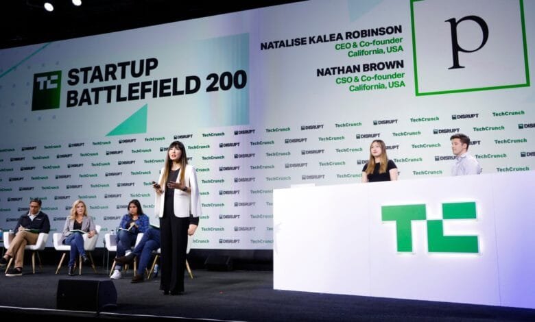 A panel discussion at TechCrunch Disrupt 2024. A woman in a white blazer speaks at a podium, while others sit on a stage.