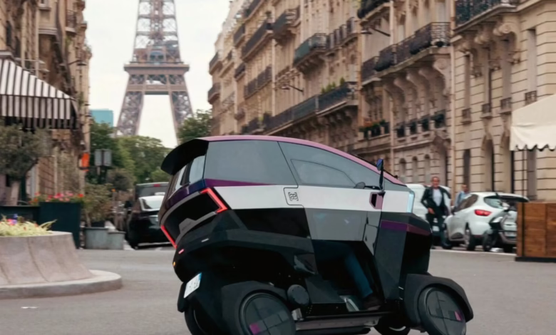 A futuristic, three-wheeled vehicle drives down a Parisian street with the Eiffel Tower in the background.