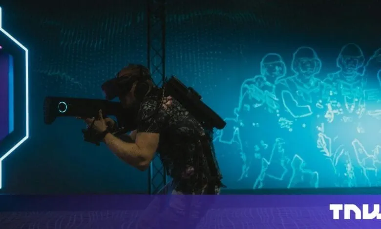 Person wearing VR headset and backpack holds futuristic weapon, immersed in a blue, digital battlefield projected on a wall.
