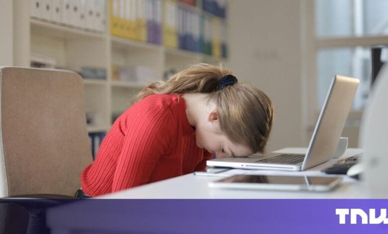 Woman in red sweater rests head on laptop, appearing stressed or overwhelmed at her desk.