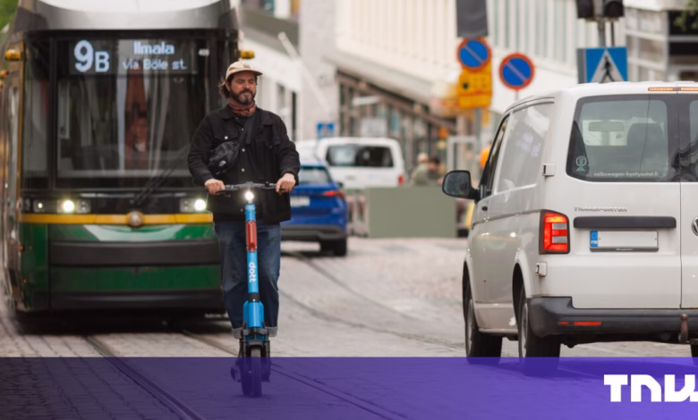 Man rides electric scooter past a tram and van on a city street.