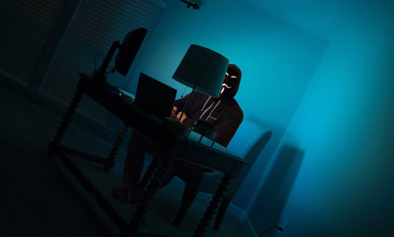 A hooded figure sits at a desk in a dimly lit room, illuminated by a lamp, working on a laptop and desktop computer. The room is painted teal.