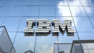 The IBM logo, rendered in metallic silver, is affixed to a modern glass building against a partly cloudy sky.