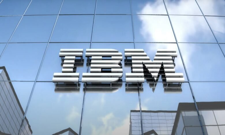The IBM logo, rendered in metallic silver, is affixed to a modern glass building against a partly cloudy sky.