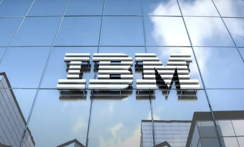 The IBM logo, rendered in metallic silver, is affixed to a modern glass building against a partly cloudy sky.