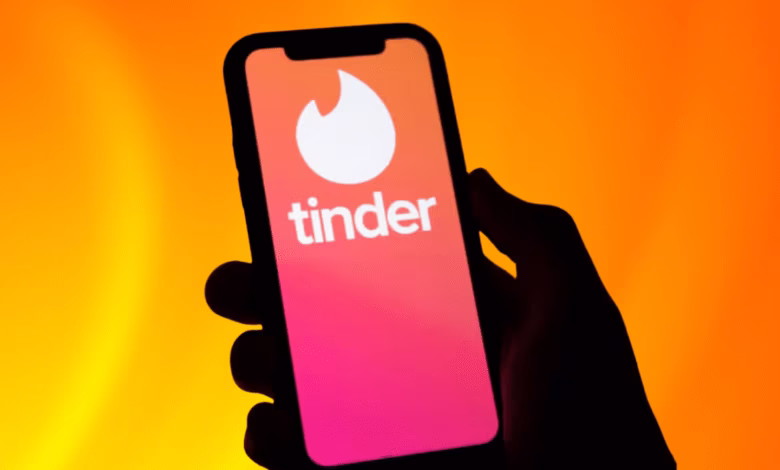 Silhouetted hand holds a smartphone displaying the Tinder app's logo and name against a vibrant orange background.