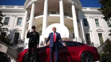 Donald Trump and Elon Musk stand before the White House with a red Tesla.