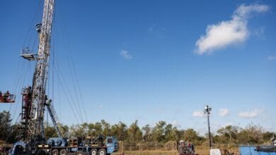 Tall oil drilling rig under a clear blue sky with a few fluffy clouds. Workers are visible on the rig and in a lift.