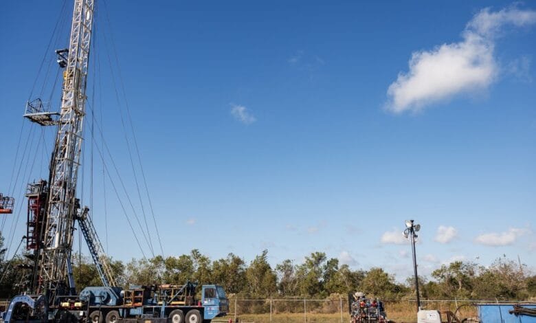 Tall oil drilling rig under a clear blue sky with a few fluffy clouds. Workers are visible on the rig and in a lift.