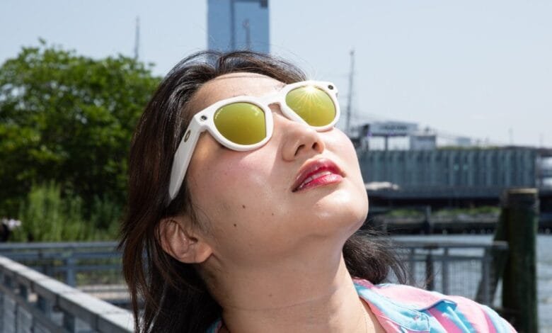 Woman wearing white sunglasses with yellow lenses, looking up towards the sun in a city setting.
