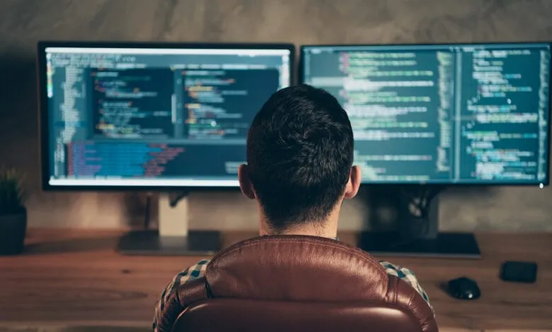 A programmer sits at a desk facing two large monitors displaying lines of code. The room is dimly lit, creating a focused atmosphere.
