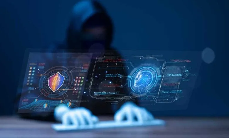 A hooded figure uses a computer displaying futuristic security interfaces with data visualizations and a digital head.