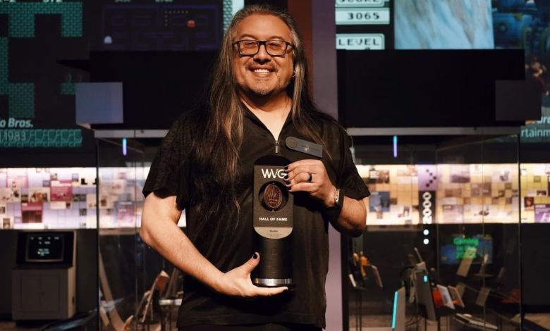 A man with long dark hair and glasses smiles, holding a World Video Game Hall of Fame award.