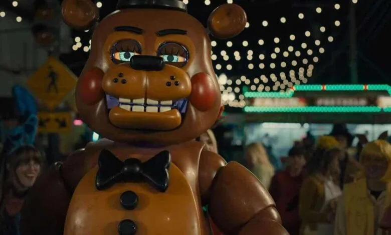 A large animatronic bear, Freddy Fazbear, stands at a nighttime carnival, surrounded by blurry figures in costumes.