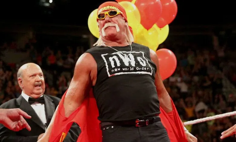 Hulk Hogan, professional wrestler, stands in a wrestling ring wearing a black nWo shirt and red cape, surrounded by balloons.