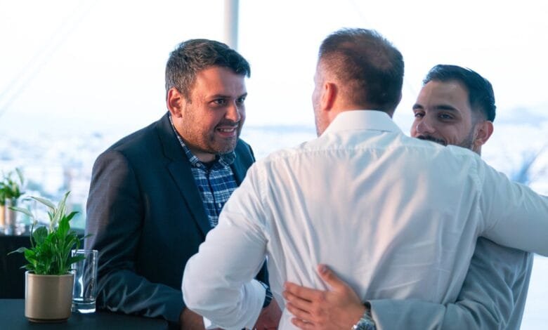 Three men in business attire embrace warmly in a bright, modern setting. One man wears a white shirt, another a dark suit.