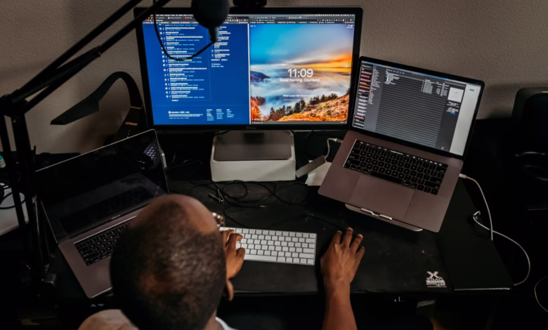 Person sits at desk working on multiple computer screens displaying code and a login screen. A microphone is visible.